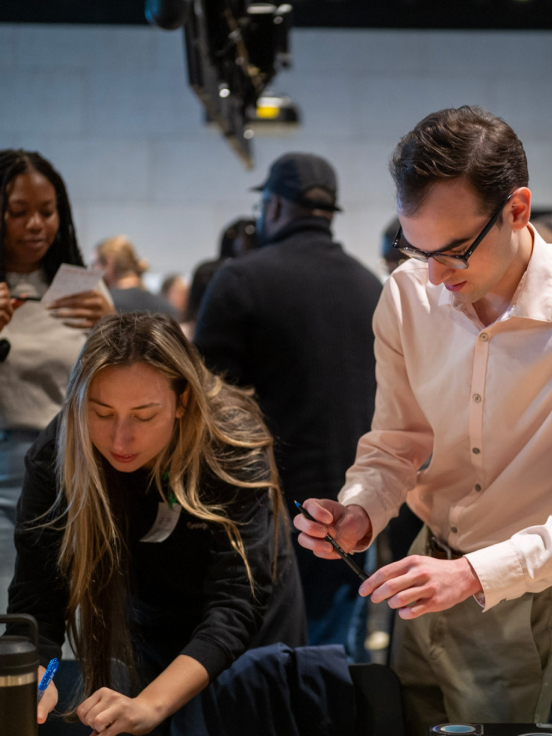 A woman is writing something, with a man standing next to her and a crowd in the background.