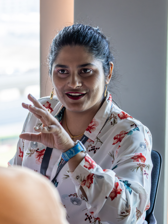 A confident-looking woman wearing a white shirt and a blue watch smiles as she engages in conversation.